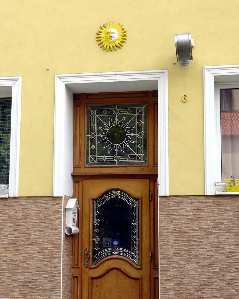 Žlutá fasáda Penzionu Slunce, dřevěné dveře s vitráží slunce a keramické slunce nad vchodem